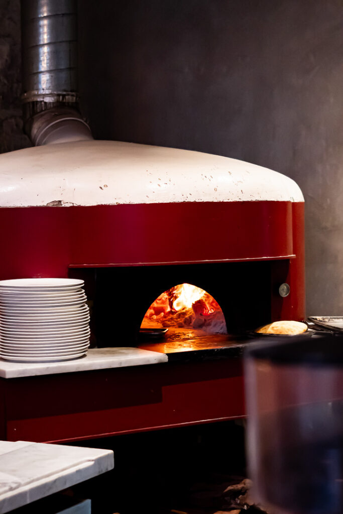 A pizza chef pulls a blistered Neapolitan pie from a glowing wood‑fired oven, embers illuminating the crust.