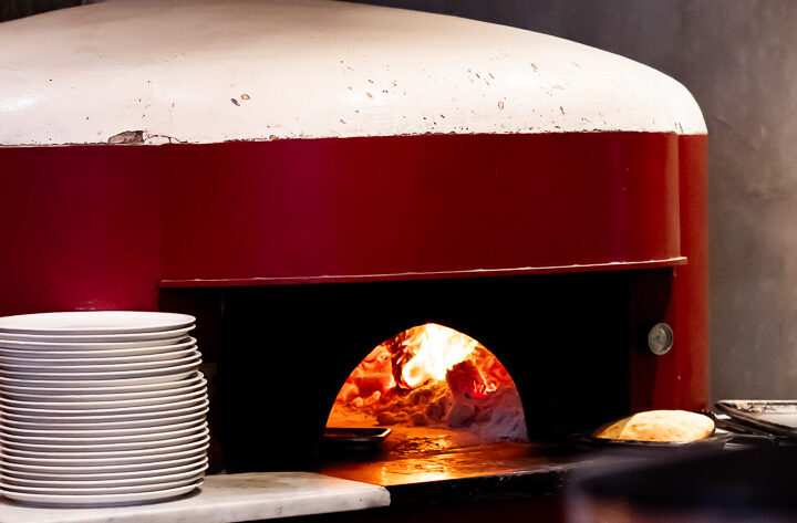 A pizza chef pulls a blistered Neapolitan pie from a glowing wood‑fired oven, embers illuminating the crust.